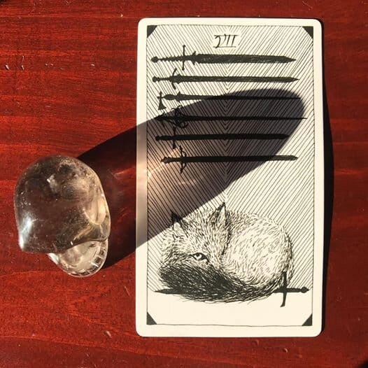 a quartz crystal skull alongside an illustrated tarot card of a fox sleeping with one eye open on the edge of a sword and 6 swords hanging horizontally above her