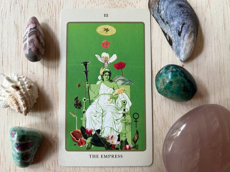 a green tarot card showing a figure seated on a throne against a light wood background surrounded by shells and stones