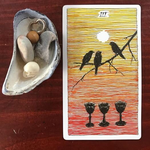 a mussel shell filled with pebbles and beads alongside a tarot card depicting 3 black goblets underneath a branch with 3 black birds and a white sun