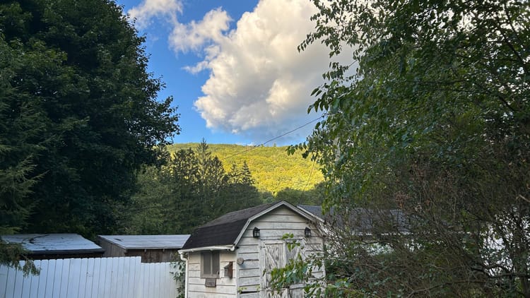 a weathered white barn in front of white fence surrounded by green trees with a sunlight-faced green mountain, blue sky and clouds in the background