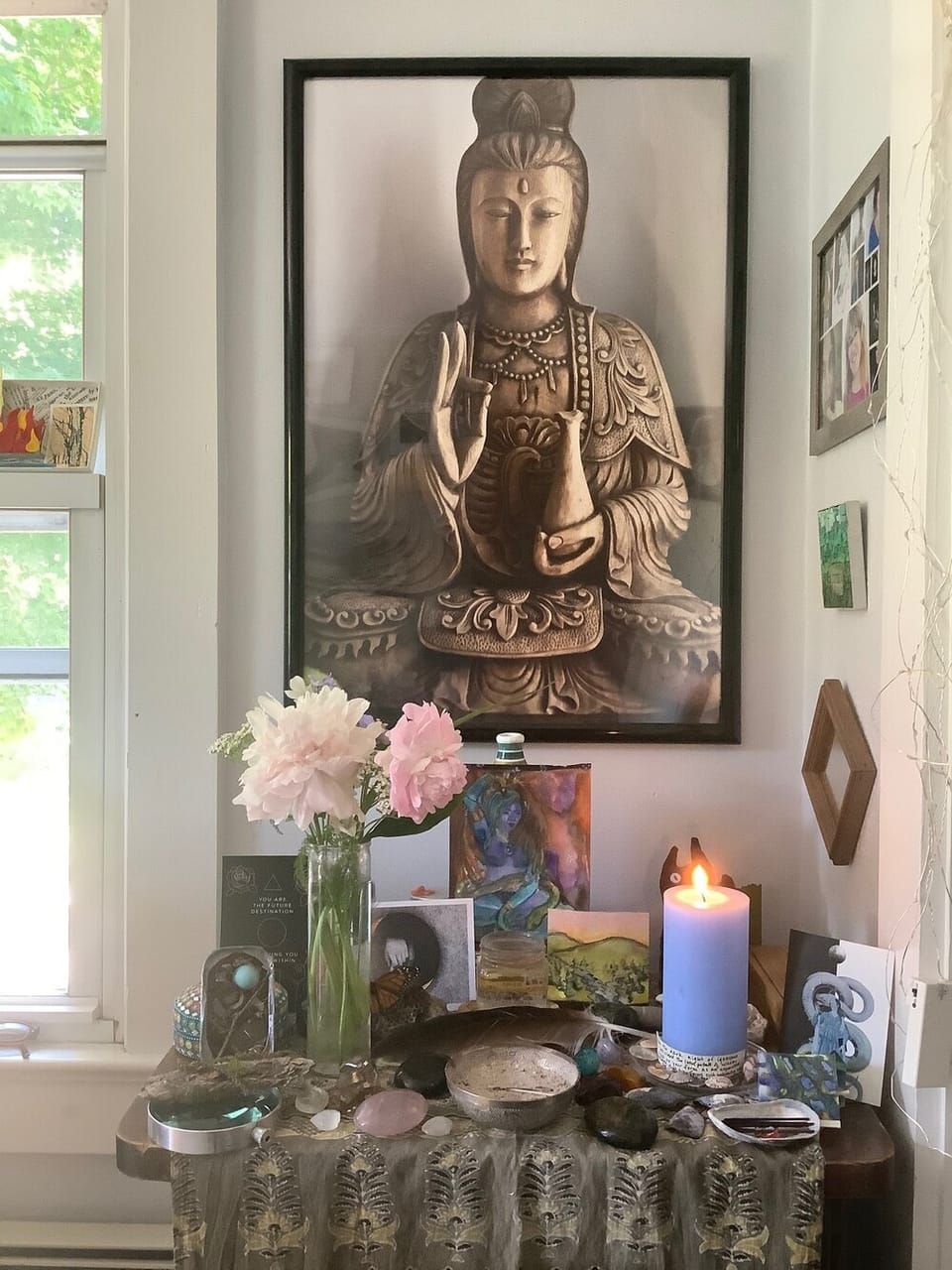 an altar with a framed image of Kannon Bodhisattva, pink peonies, a lit blue candle, an incense bowl and various stones, feathers and sundry objects
