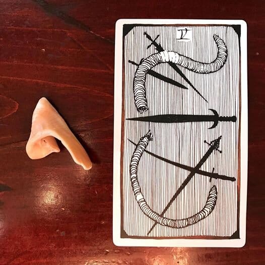 a seashell fragment next to an illustrated tarot card of a severed earthworm, 5 black swords an a background of thin vertical lines