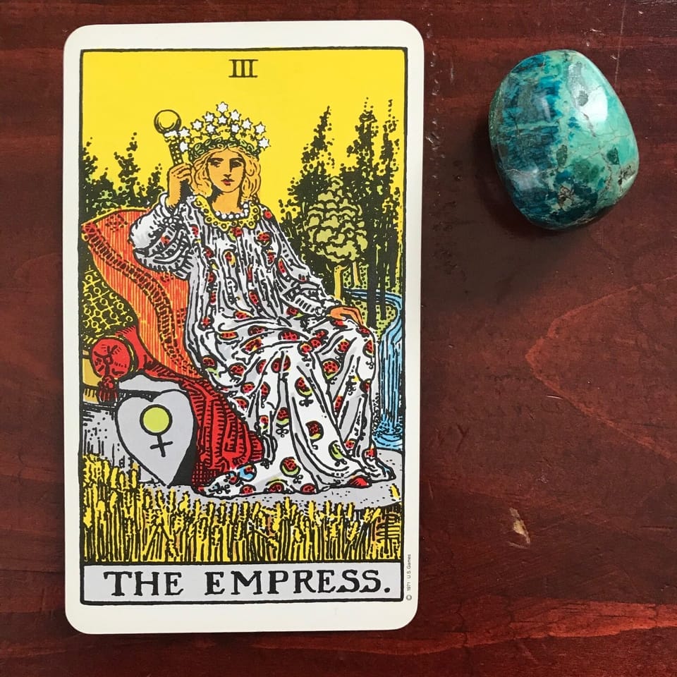 a tarot card depicting a white femme figure in a pomegranate mumu holding a scepter and a turquoise stone against a dark wood background