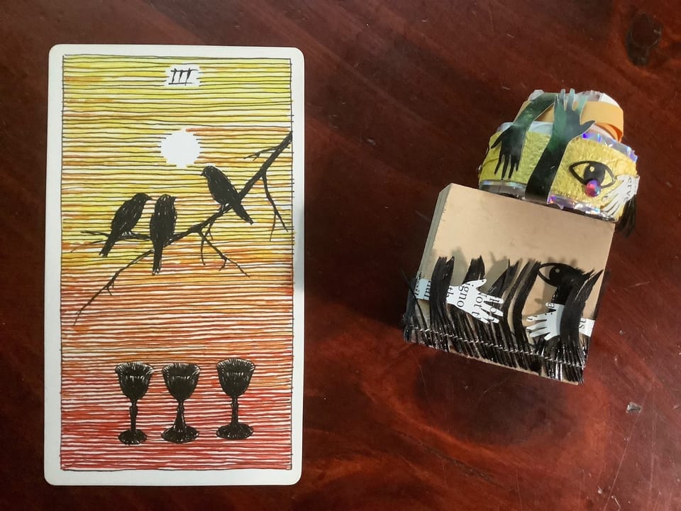a yellow-orange tarot card with three black birds and three black cups and a small sculptural artwork against a dark wood background