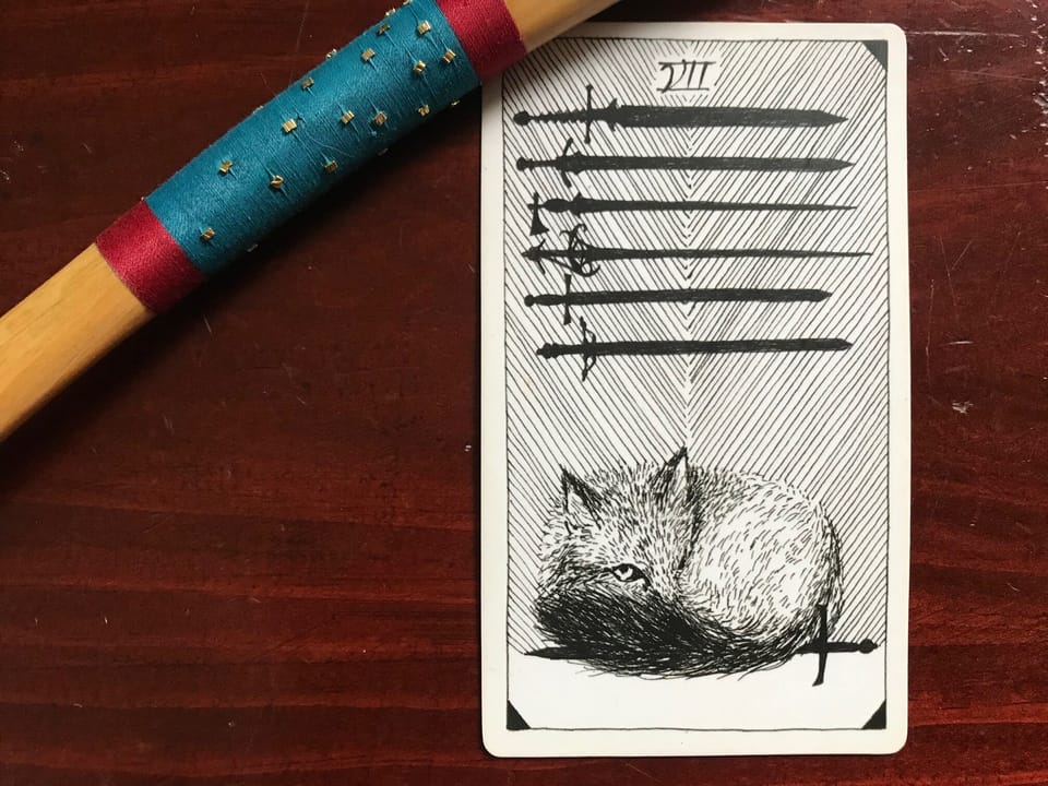 a tarot card of a fox sitting on a sword with 6 swords above and a segment of a pine wood wand wrapped in red and blue thread against a dark wood background