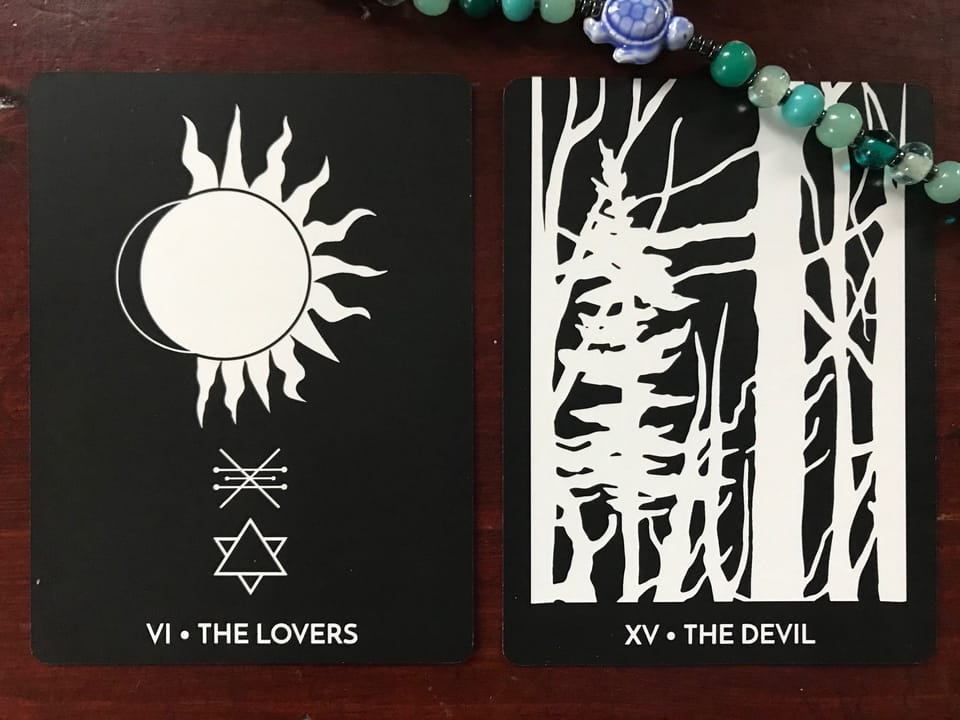 two boldly graphic black and white tarot cards against a dark wood background with green round beads and a blue turtle bead in the upper corner