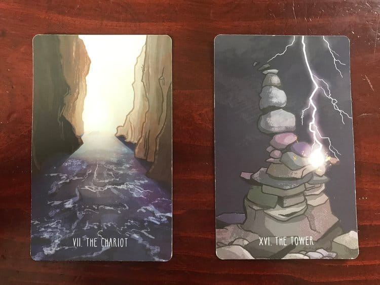 2 tarot cards against a dark wood background, one showing a river running through a canyon, the other showing a bolt of lightning striking a cairn