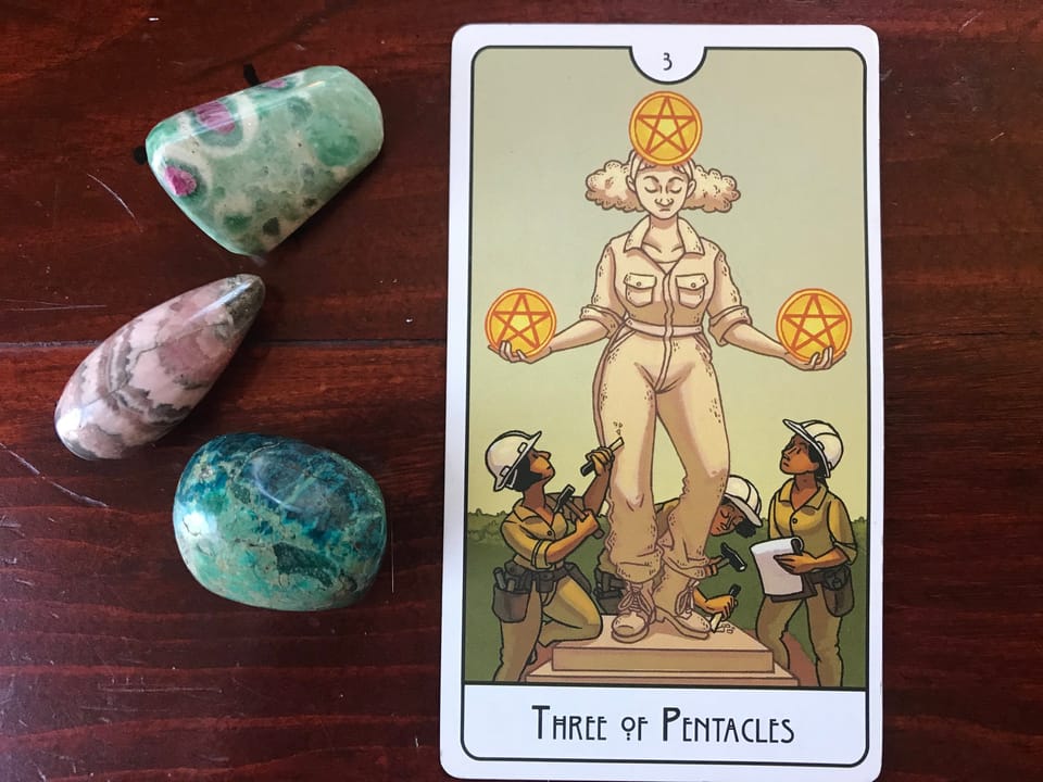 a tarot card showing 3 figures working on a statue holding 3 gold pentacles, against a dark wood background with 3 colored stones