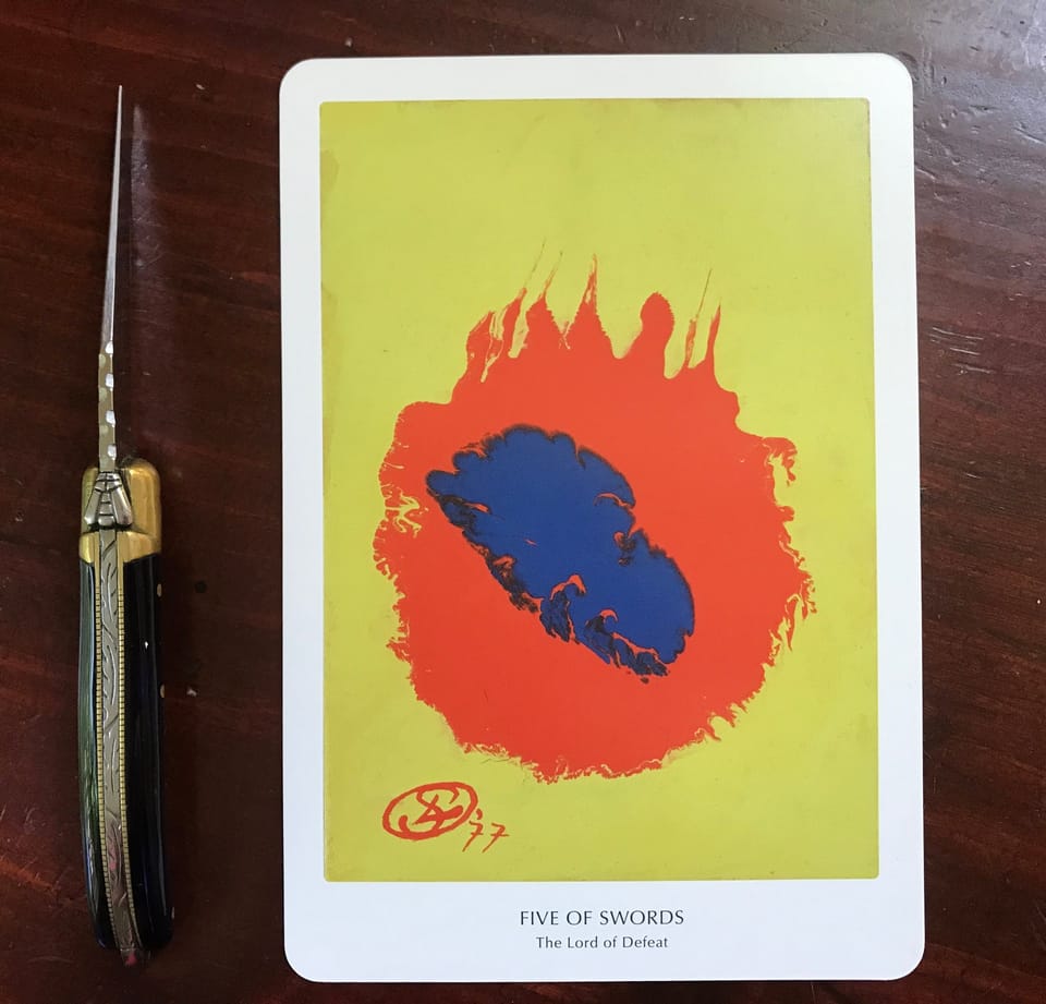 an abstract yellow, red and blue tarot card against a dark wood background with an extended pocket knife to the side on which there is a silver bee