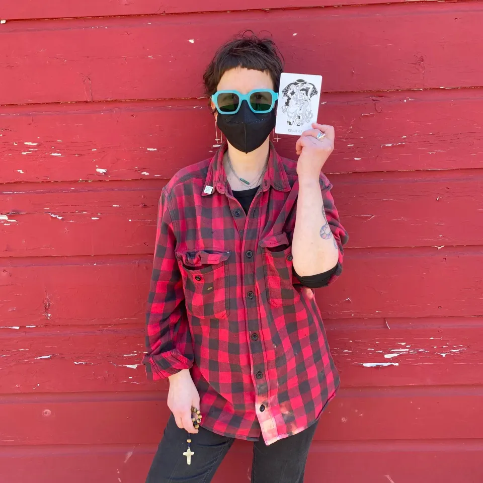a white person with short brown bangs, turquoise sunglasses and a black and red plaid shirt holding a rosary in one hand and a tarot card in the other standing against a red barn