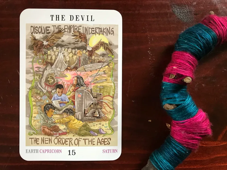 a tarot card and a blue and pink string-wrapped crooked stick against a dark wood background